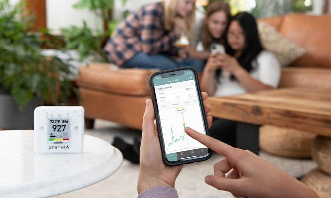 Indoor CO2 monitor on a table to the left, with the phone readings focus in with the air readings with people in the background